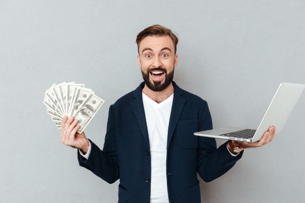 happy-surprised-bearded-man-holding-money-laptop-computer-while-looking-camera