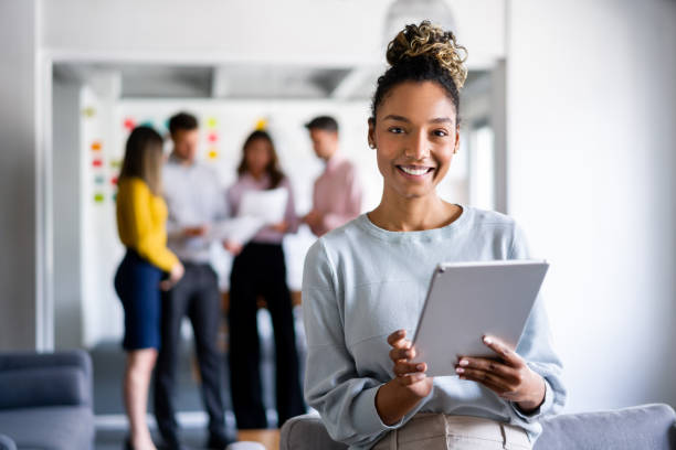 Women with people behind her,she is holding a tablet and smiling