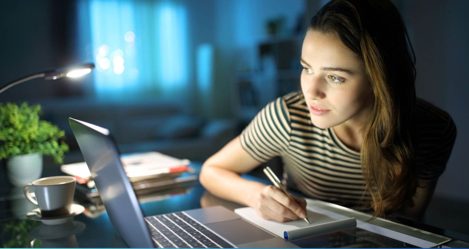 GIRL WRITING ON A NOTEBOOK LOOKING AT HER LAPTOP SITTING AT DESKDESK